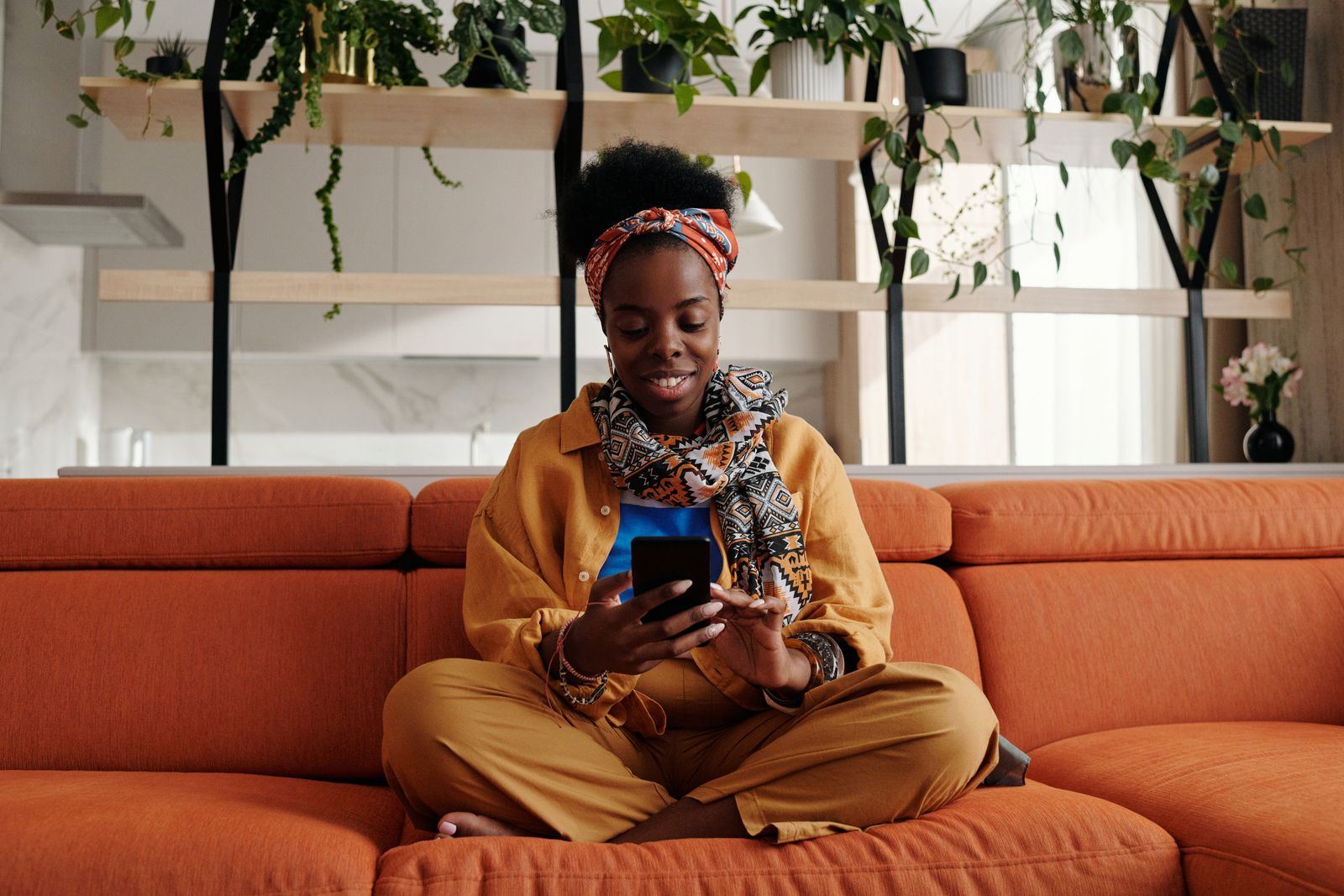 Une femme au bandeau Ankara sourit à son téléphone sur un canapé terracotta